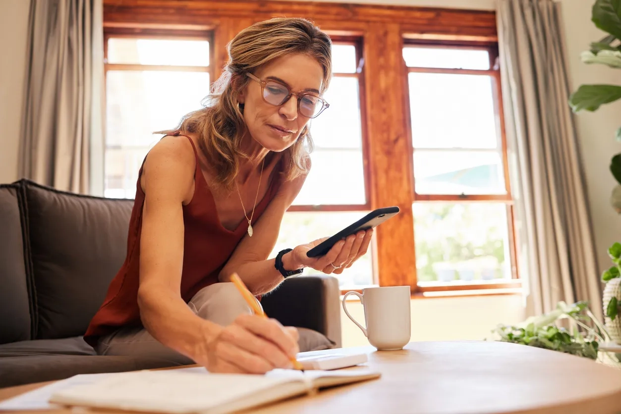 woman 60s holding phone while writing notes in a book