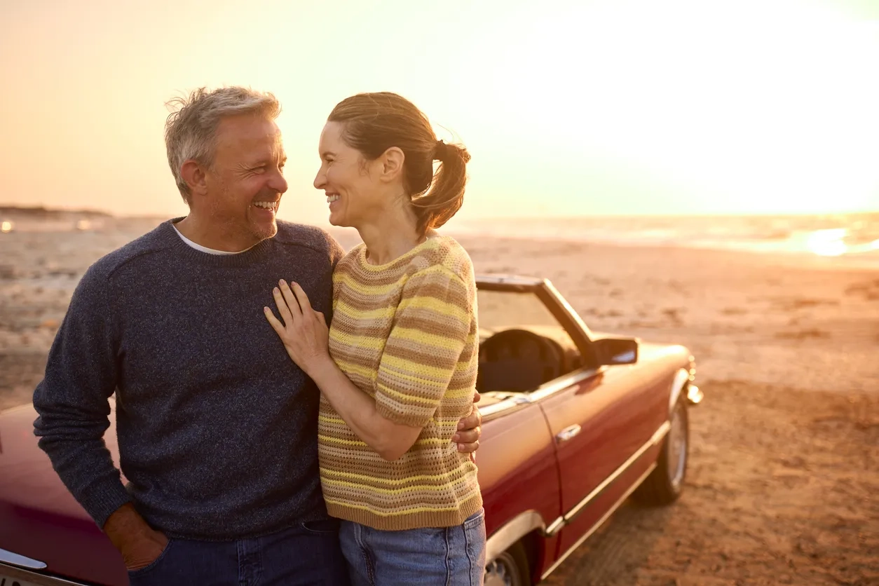 Loving Retired Senior Couple On Vacation Next To Classic Sports Car At Beach Watching Sunrise