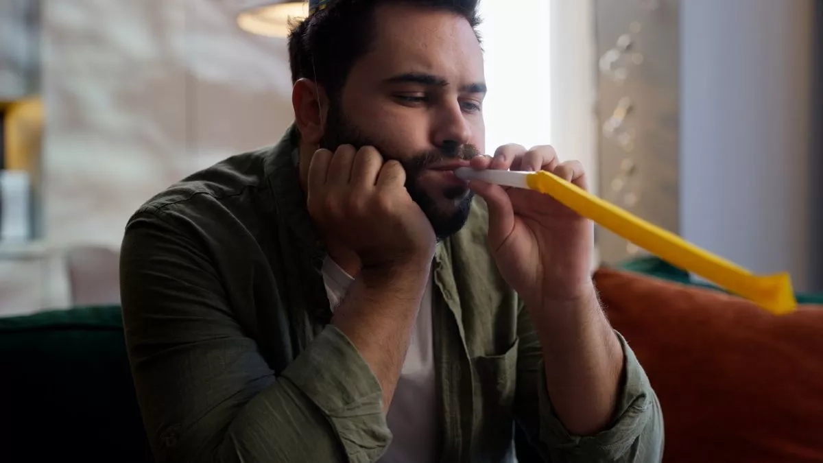 Lonely sad upset depressed despair man in festive cap on head blowing in whistle celebrating birthday alone