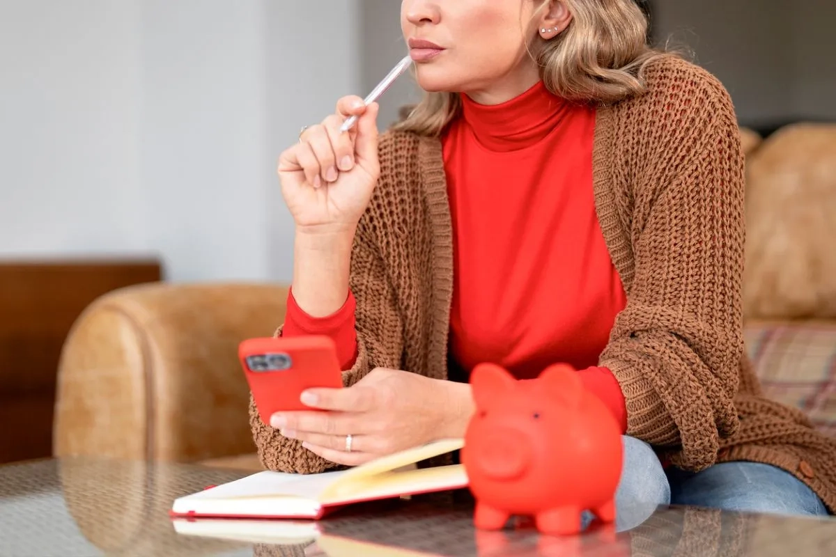Woman sits and thinks while holding a phone and pen with a piggy bank nearby in a cozy space