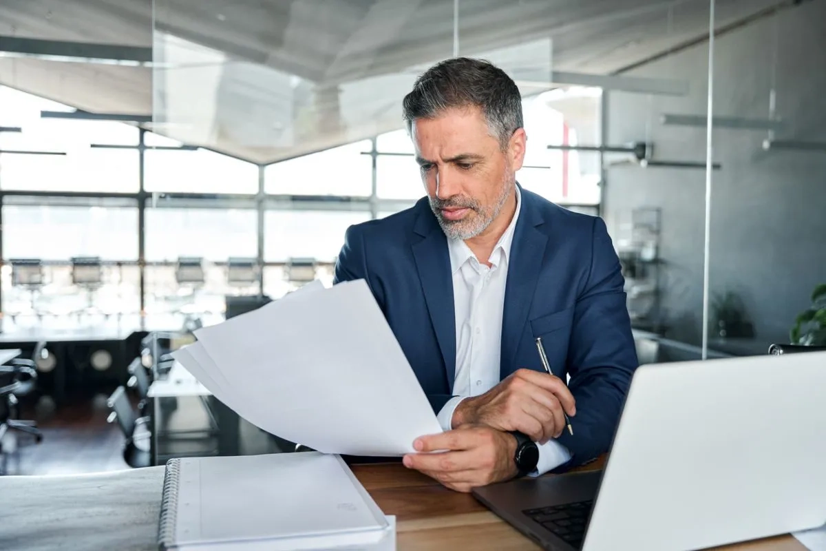 Serious busy middle aged professional business man executive ceo wearing suit sitting at desk in office working checking bills corporate financial accounting documents.