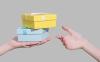 Two gift boxes of blue and yellow color in hand on gray background give in other hand.